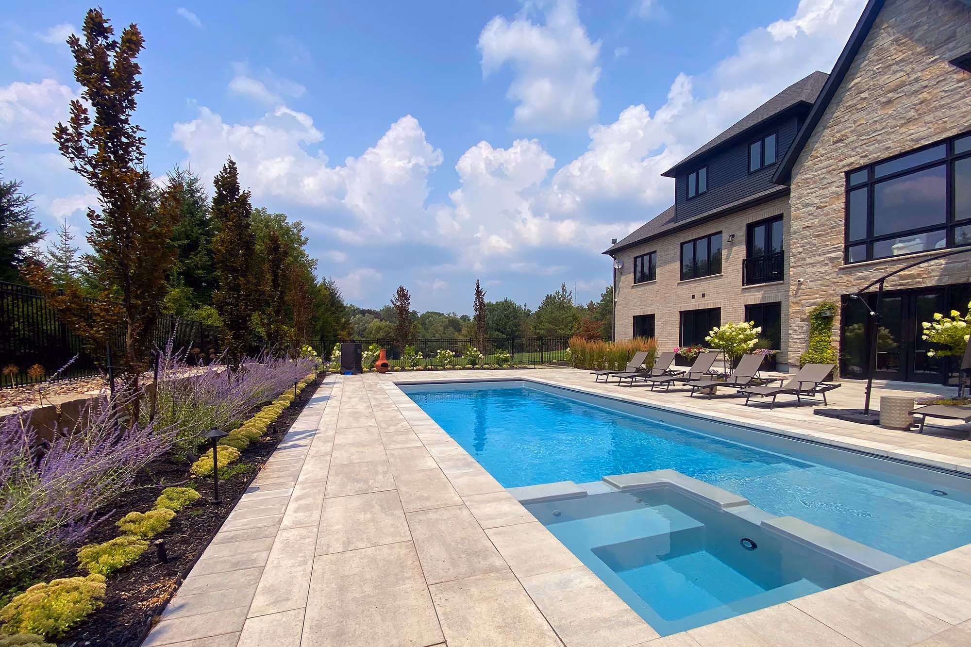 Pool with visible spa steps, stone patio and landscaped perennial beds beside large home
