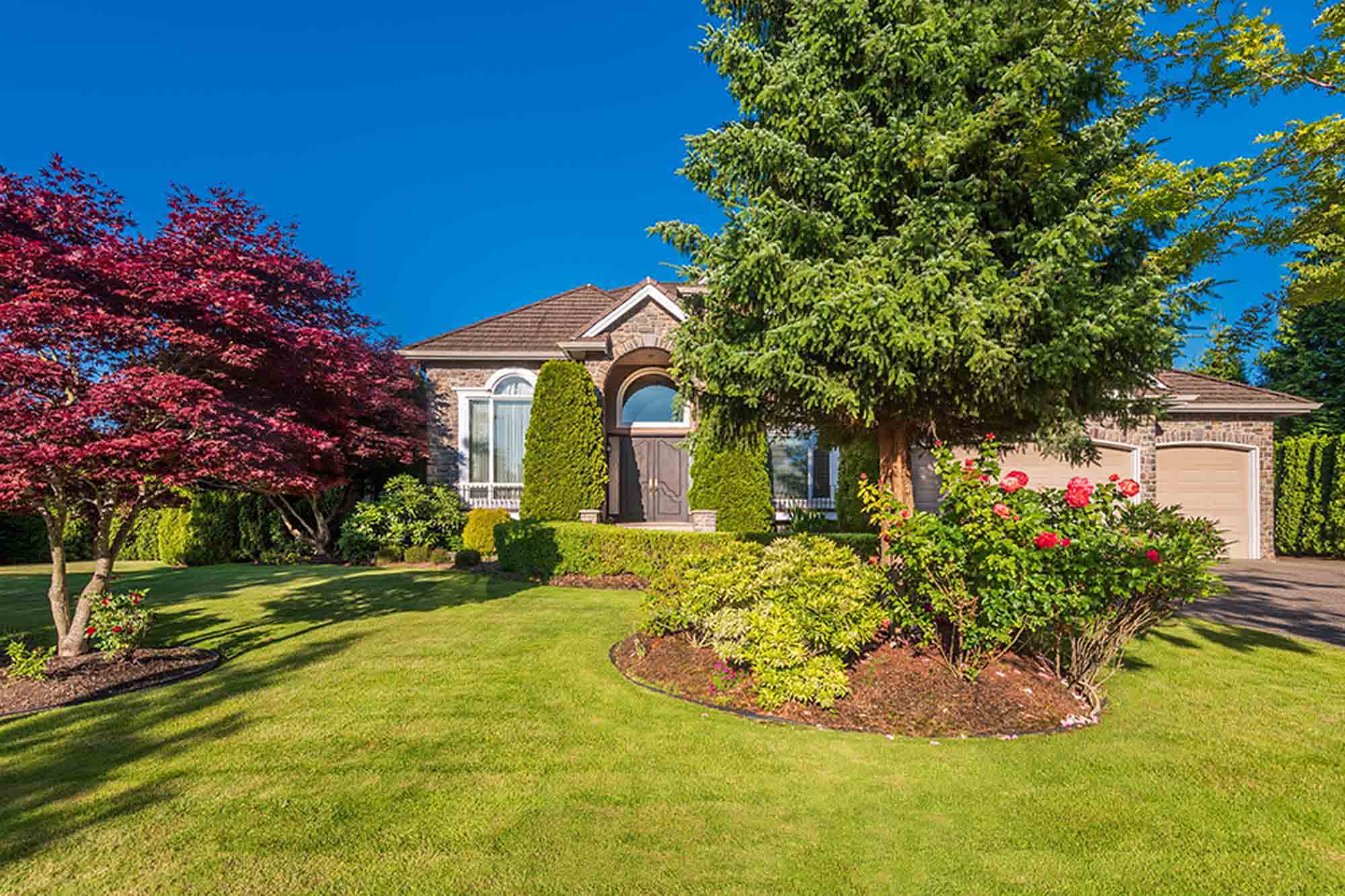Well-maintained residential front yard with manicured lawn, curved planting beds, ornamental trees, and a brick home entrance in Ontario