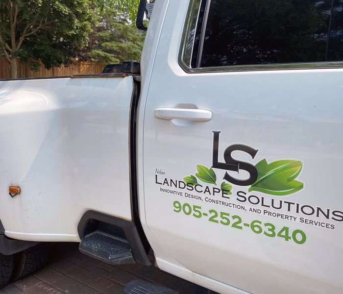 White work truck door with a "LS Landscape Solutions" logo, green leaf graphic and phone number printed on the side, parked on a residential driveway.